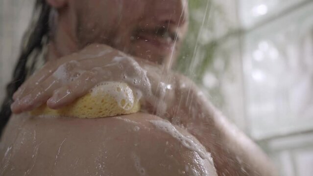 Caucasian long haired man cleaning her skin sponge relaxing in shower from shoulder to face. Close-up European adult beard male using soap for daily hygiene. Personal body care in bathroom at home. 