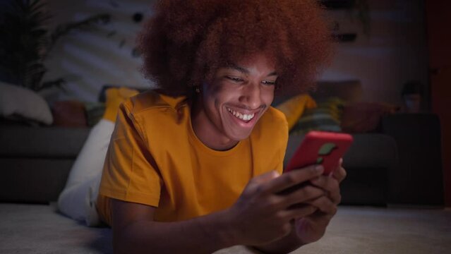 Close up of cheerful young afro boy using smart phone lying on carpet in living room. Smiling man chatting on mobile at home at night. Single people, dating apps and social networks in generation z.