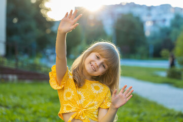 Preteen child kid smiling friendly at camera, waving hands gesturing hello, hi, greeting or goodbye, welcoming with happy expression. Pretty blonde girl standing on city sunset park street, outdoors