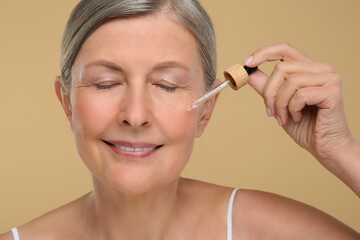 Senior woman applying cosmetic product on her aging skin against beige background. Rejuvenation treatment