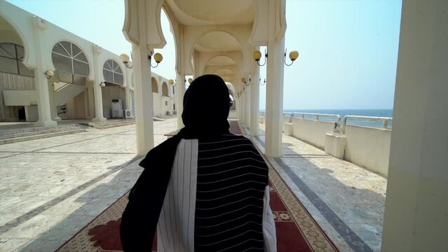 A woman in black and white arabic clothing walking on a carpet in a place with arches, in a mosque near the Red Sea in Jeddah, Saudi Arabia. 