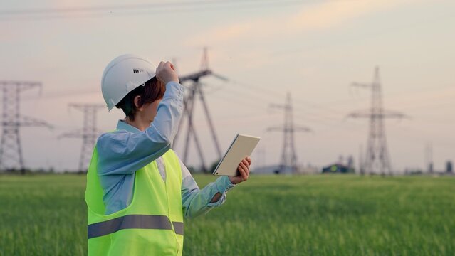 Energy security concept, ecology. Civil engineer, woman specializing in electricity supply works outdoors. Modern technologies. Power engineer in safety helmet checks power line, digital tablet