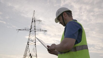 Electrician checkups power transmission lines with tablet standing near tower
