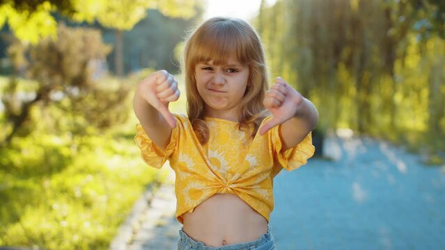 Dislike. Upset preteen child kid showing thumbs down sign gesture, expressing discontent disapproval dissatisfied bad work mistake. Displeased blonde girl standing on city sunset park street, outdoors