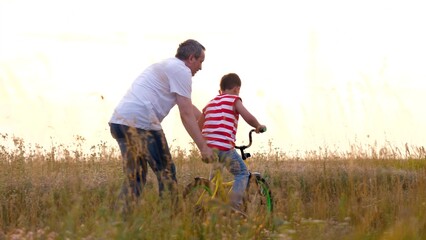 Father holds bike and little son trains to ride vehicle at countryside at sunset