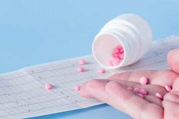 A hand pours a handful of pink pills from a white jar onto an electrocardiogram of the heart, on a...