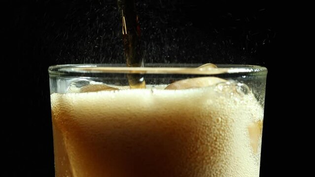 Close-up shot of cola pouring into a glass with ice cubes splashed with soda bubbles in it.