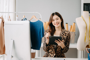 Successful fashion designer. Attractive young asian woman with smile while standing in workshop. Beautiful owner business woman working and holds tablet, laptop and smartphone on desk in studio..