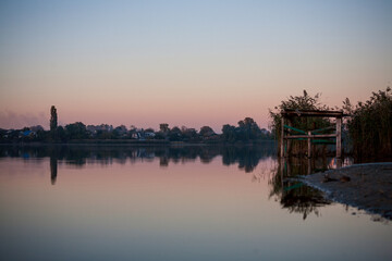 Autumn landscape on the river at dawn, where the sun is rising on the horizon and the sky is pink and blue with white clouds and a bridge, and in the distance reeds and trees