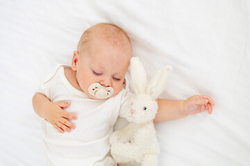 cute little newborn baby sleeping with a pacifier in his mouth under a blanket hugging a plush bunny, sweet healthy baby sleep in a white crib