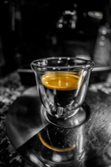 Glass cup with freshly brewed black coffee with foam and golden cream with visible bubbles on top of a metal coffee plate with its reflection of the coffee foam. Dark black and white background.