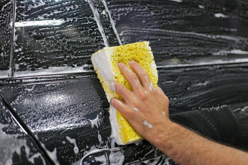 Car detailing series: Close-up of hand cleaning black car of serviceman. The man using yellow sponge to wash his car. The process of washing cars with foam and sponge.