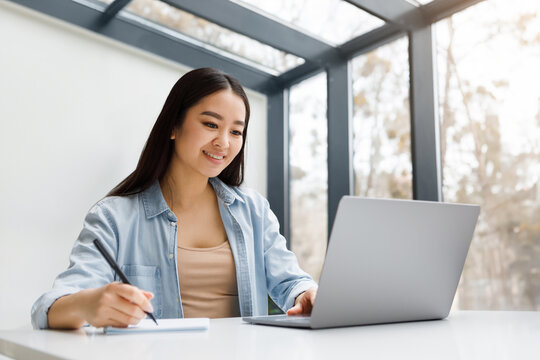 Positive busy hardworking asian student lady writing notes, webinar summary, studying in college library with laptop