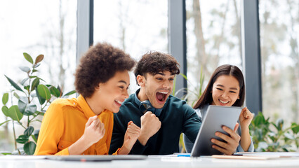 Overjoyed multiracial classmates looking at digital tablet screen, shouting yes and clenching fists...