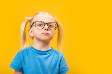 Portrait of little blonde girl with two tails wears glasses and looks pensively into the distance. Preschooler on yellow background.