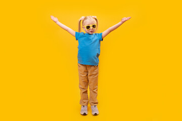 Happy little girl in blue T-shirt and sunglasses raised her hands up. Portrait of preschooler on yellow background.
