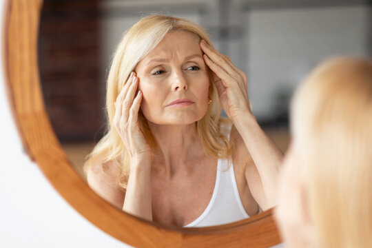 Upset middle aged woman looking at mirror and touching wrinkles on her face, lady examining fine lines on forehead