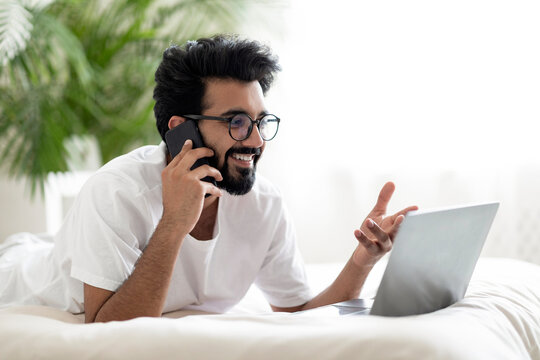 Indian man talking on cellphone and using laptop while relaxing in bed - Powered by Adobe