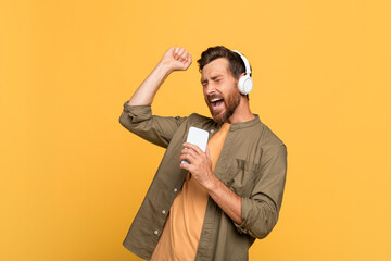 Excited middle aged man in headphones with open mouth singing at smartphone, having fun on yellow...