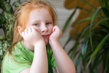 A red-haired little girl in a green T-shirt sits near the window at home and smiles. 