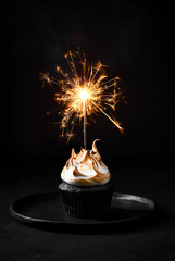 Single chocolate cupcake with meringue frosting. Sparkler in the cupcake. Black background, plate...