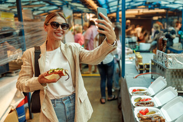 Smiling mature woman in eyeglasses make a selfie on phone of takeaway fast food on food market