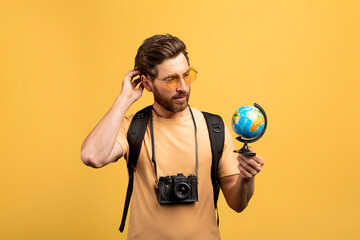 Thoughtful middle aged male traveler holding small globe, looking and thinking where to go next, yellow background