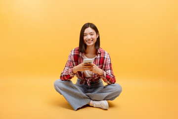 Happy asian lady using cellphone texting or browsing internet sitting on floor over yellow background, smiling at camera