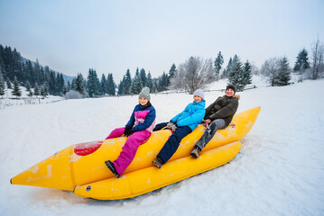 Happy family slides through the snow on an inflatable sled in shape of banana. Inflatable sled for three. Active outdoor sports in winter.