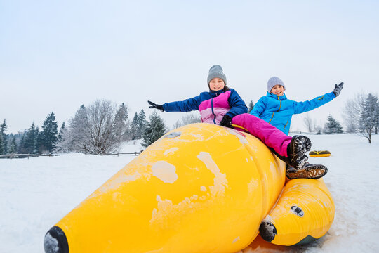 Happy children race on inflatable banana-shaped sleds in snow. Winter active outdoor entertainment.