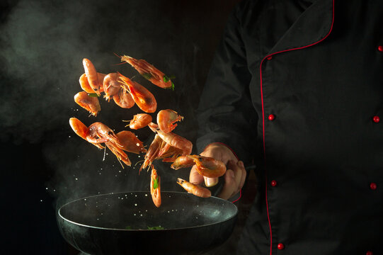The chef cooks shrimp in a hot pan with steam. The concept of cooking seafood or healthy vegetarian food and eating on a black background. Freeze in motion. Free space for recipe
