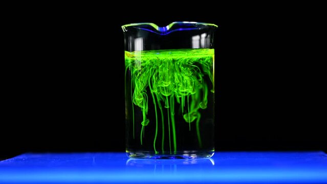 A woman scientist experimenting with a green fluorescent solution in a glass conical flask in dark biomedical chemistry laboratory for health care drug development. 4K copyspace.