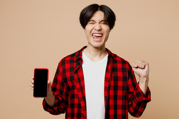 Young man of Asian ethnicity wear red shirt casual clothes hold in hand use mobile cell phone with blank screen workspace area do winner gesture isolated on plain pastel light beige background studio.