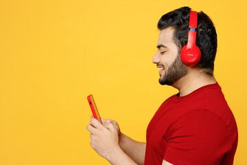 Side profile view young happy Indian man he wears red t-shirt casual clothes listen to music in headphones use mobile cell phone isolated on plain yellow orange background studio. Lifestyle concept.