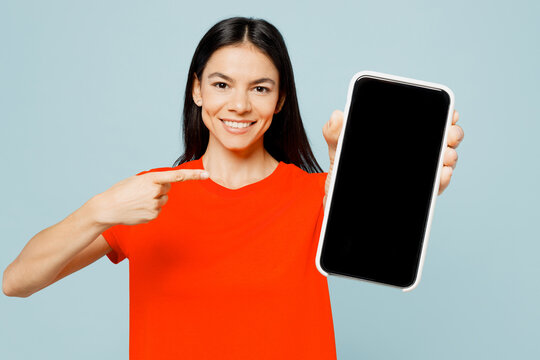 Young Latin Woman Wear Orange Red T-shirt Casual Clothes Hold In Hand Use Point Finger On Mobile Cell Phone With Blank Screen Workspace Area Isolated On Plain Blue Background Studio Lifestyle Concept