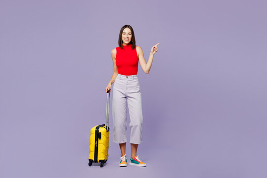 Traveler woman in red casual clothes hold suitcase point finger aside isolated on plain purple background studio Tourist travel abroad in free spare time rest getaway Air flight trip journey concept