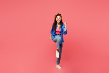 Full body young woman of African American ethnicity wear blue shirt casual clothes doing winner gesture celebrate clenching fists say yes raise up leg isolated on plain pastel pink background studio.
