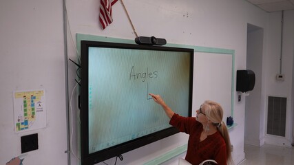 Happy female teacher writing on an interactive whiteboard teaching geometry math in a school classroom with US American flag in background.