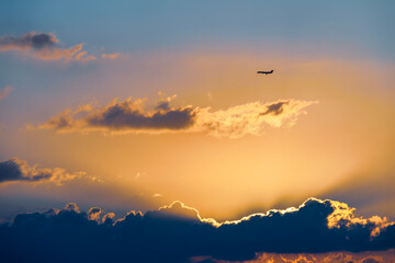 A plane flying in the sky at sunset. The plane is silhouetted against the fiery sky. The sun is setting in the distance, and its rays are visible through the clouds.