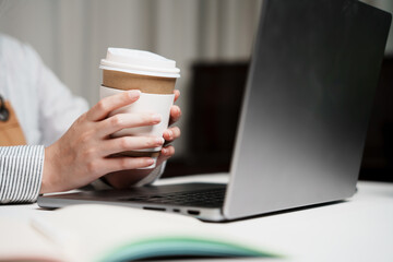 Two hands holding drinking coffee cup with screen laptop computer open for mockup multimedia technology device with plants on table indoor workspace working meeting online at home or shop cozy relax