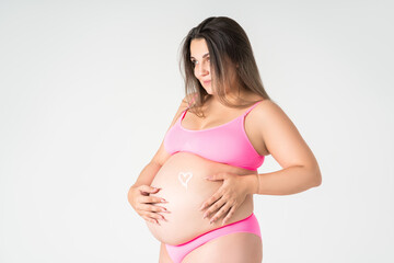 Happy plus size pregnant woman in pink underwear applying skin stretch mark cream on her belly on gray background, maternity clothes, pregnancy concept
