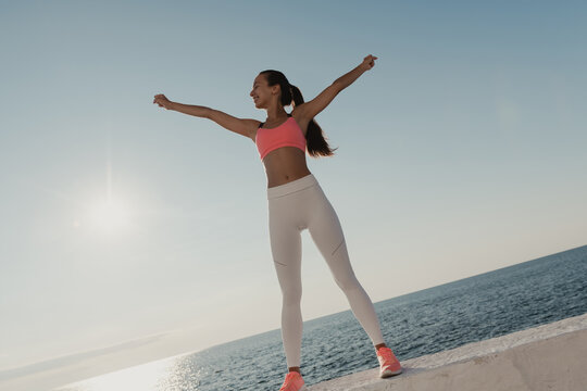 Attractive young woman in sportswear stretching out hands and looking happy while standing seaside