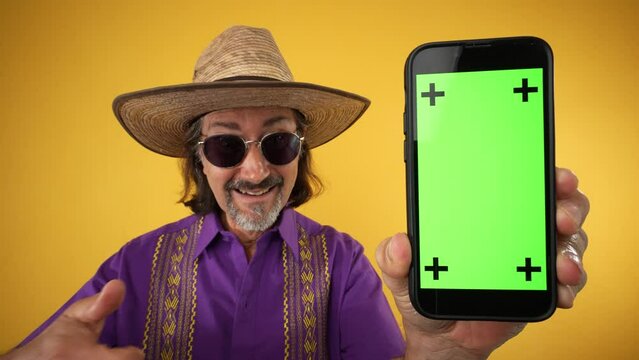 Portrait of happy mature elderly hispanic man wear ethnic shirt hold in hand point finger on mobile cell phone with blank green screen tracking points on solid yellow background studio