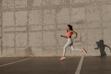 Beautiful young woman in sportswear running in front of the concrete wall outdoors