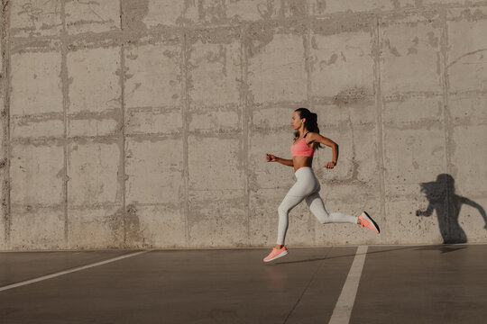 Beautiful young woman in sportswear running in front of the concrete wall outdoors