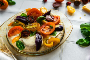 Step by step process of making colorful vegetable and mozzarella  focaccia bread. Focaccia topped with variety of tomatoes, peppers and mozzarella slices in glass baking form on white surface.