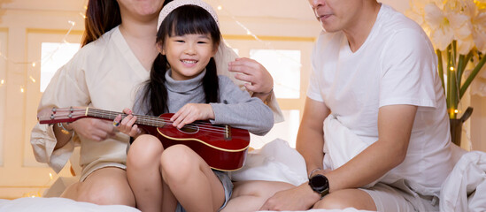 Happy asian family father and mother sitting teaching play ukulele guitar with daughter together on bed in bedroom at home, mom and dad relax with learning music with kid, lifestyles concept.