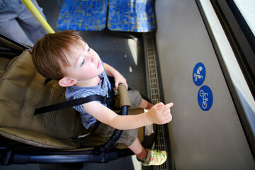 The toddler is sitting in a stroller on the bus, with people walking by outside. Kid boy aged two years (two-year-old)
