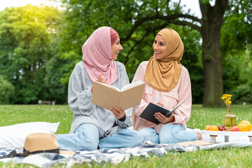 Two beautiful Arab women in hijabs are sitting on a blanked in the park. Smiling friends at a...