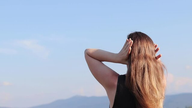 Girl tourist straightens her hair, hot sunny day. Girl against the blue sky. A woman in nature breathes fresh air, rear view. Hair with a crease from an elastic band